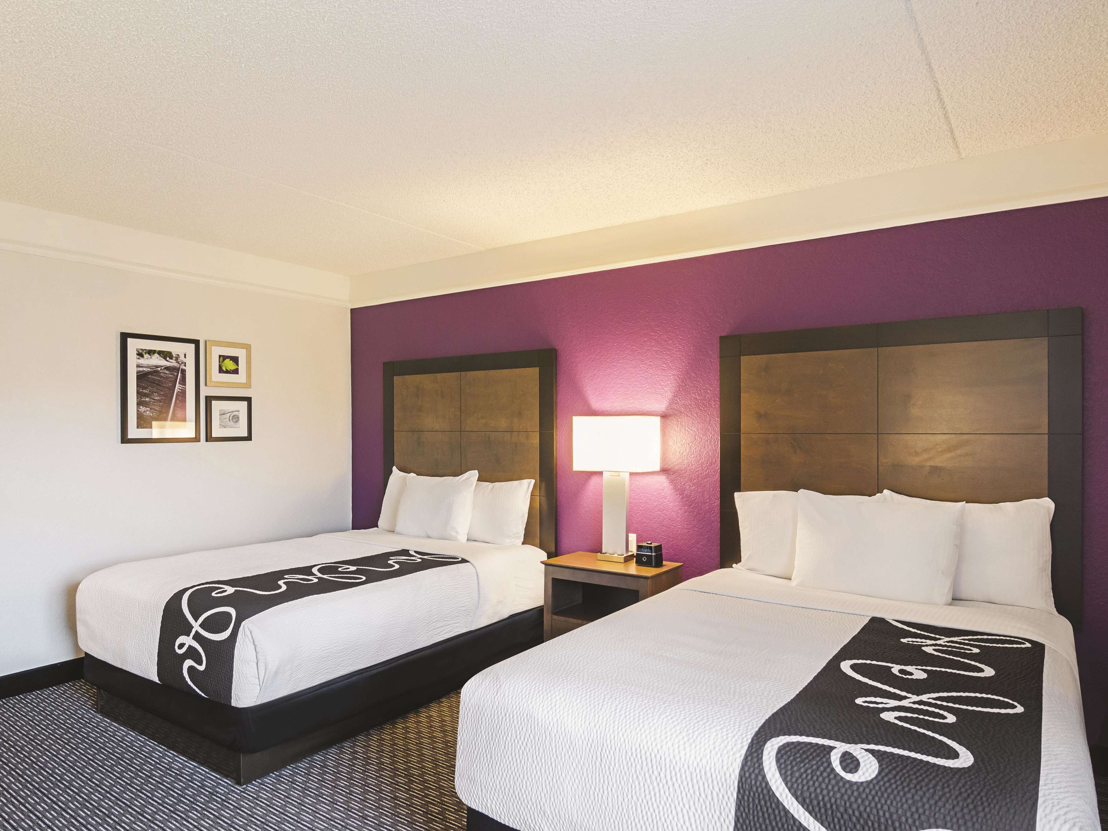 Habitación de hotel con dos camas, una mesita de noche con lámpara y una pared decorada con cuadros.