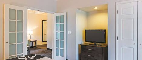 Quarto de hotel com cama, televisão em penteadeira e porta que dá para outro cômodo.