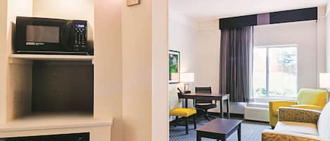 A hotel room with a microwave on a shelf, a dining table with chairs, a sofa, and a large window with curtains.