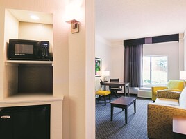 Habitación de hotel con microondas en un estante, una mesa de comedor con sillas, un sofá y un ventanal con cortinas.