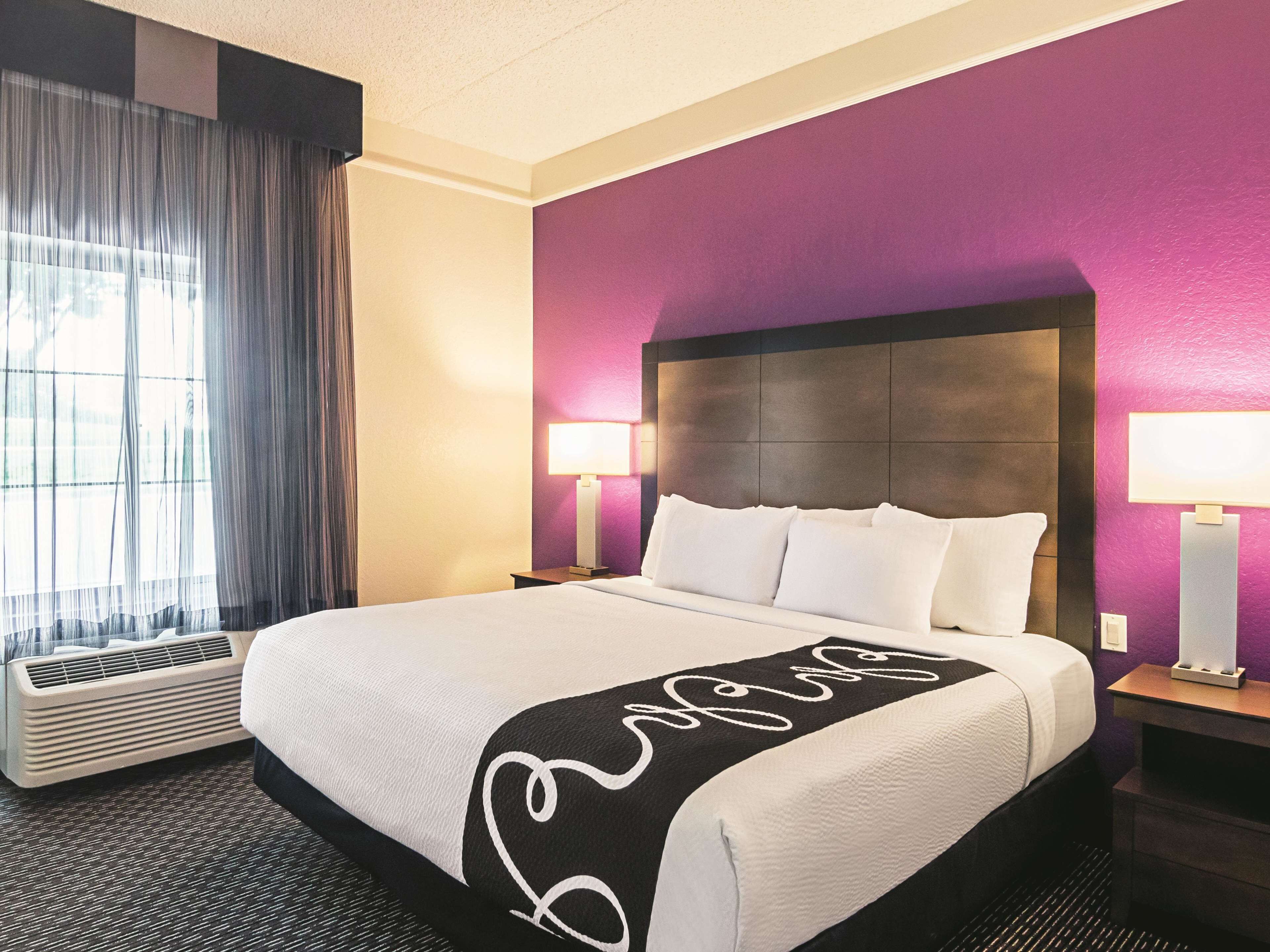 A hotel room with a large bed, two bedside tables with lamps, a window with curtains, and a wall heater.