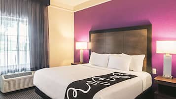 A hotel room with a large bed, two bedside tables with lamps, a window with curtains, and a wall heater.