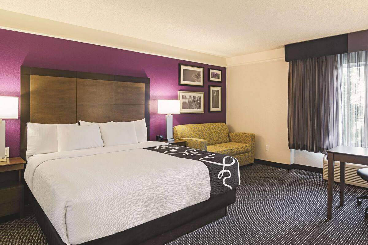 Habitación de hotel con una cama grande, un sofá pequeño, un escritorio y un ventanal con cortinas.
