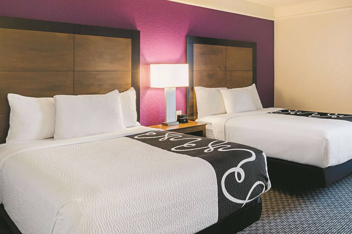 Habitación de hotel con dos camas, una mesita de noche, una lámpara y un cabecero con textura.