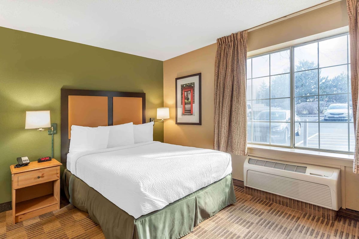 Une chambre d’hôtel avec un lit, des lampes de chevet, une table de chevet, une fenêtre avec des rideaux et un tableau encadré au mur.