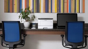 A workspace with two blue office chairs, a printer, a laptop, a potted plant, and a colorful striped wall art.