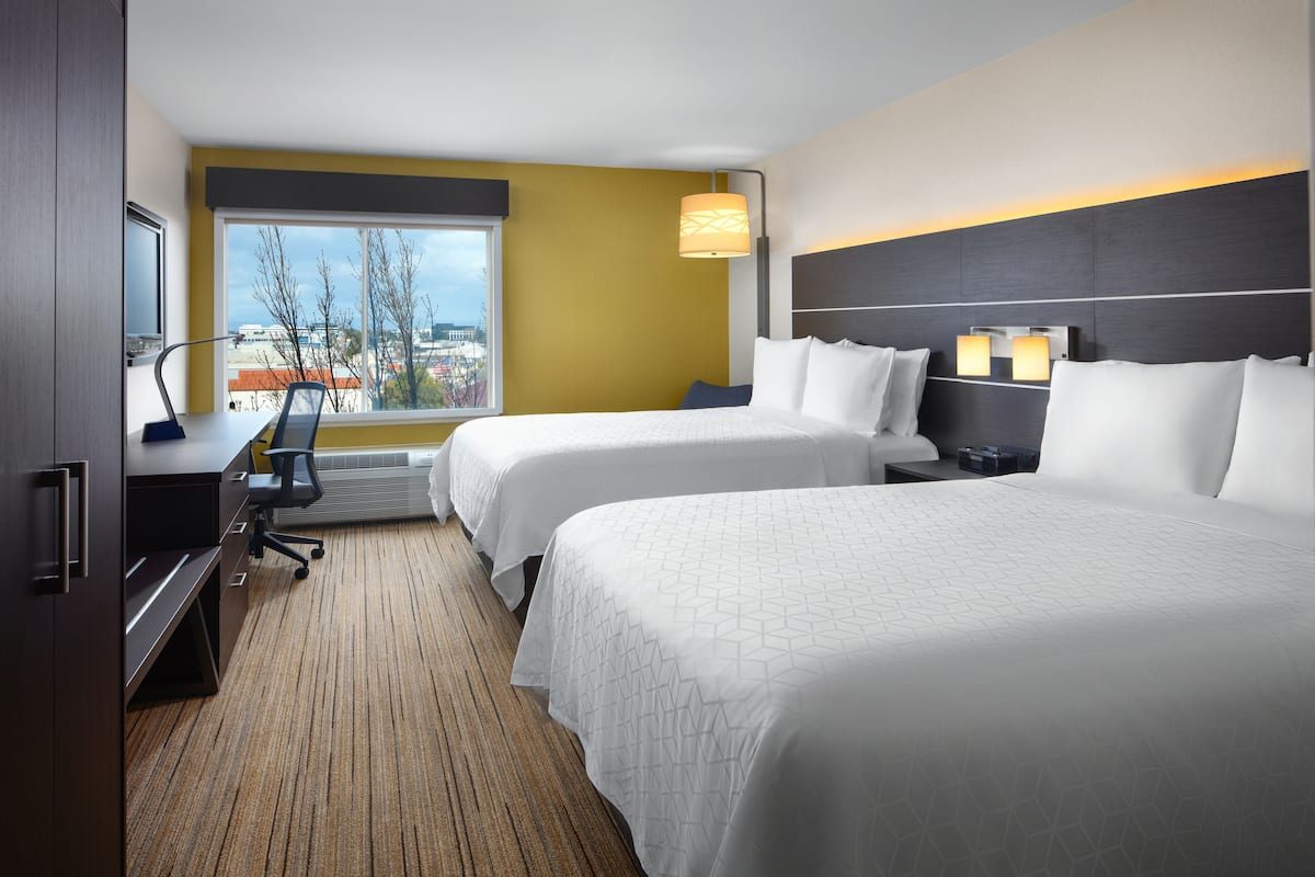 A hotel room with two beds, a desk, a chair, and a large window offering a city view.
