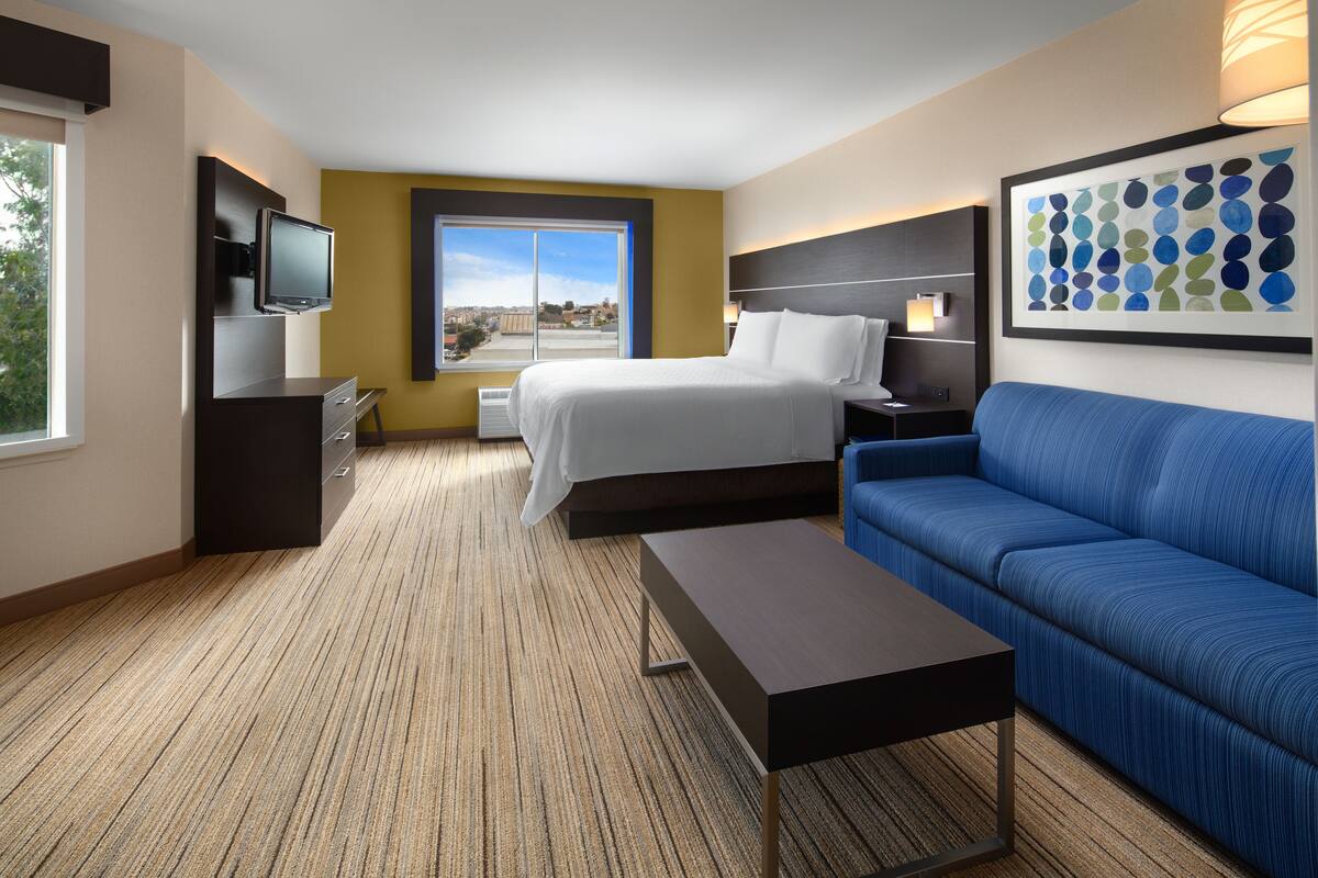 A hotel room with a bed, a TV, a sofa, and a large window with a city view.