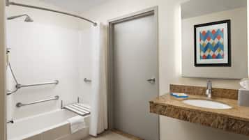 A modern bathroom with a walk-in shower, grab bars, a white bathtub, a granite countertop sink, and a framed artwork above the mirror.