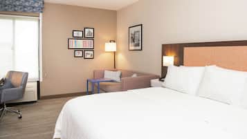 A hotel room with a bed, a desk, a chair, and a sofa. There are framed pictures on the wall and a lamp on the desk.