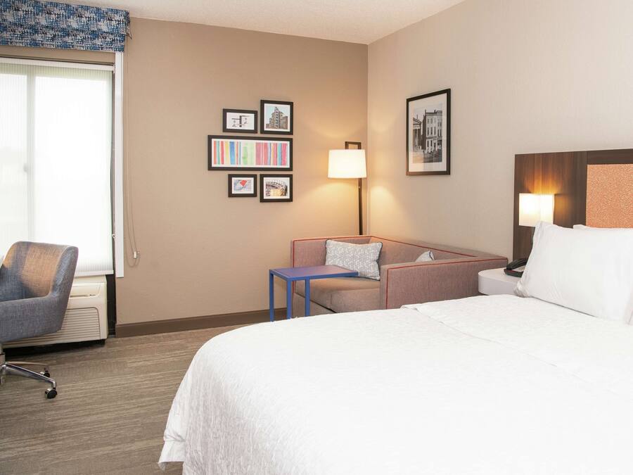 A hotel room with a bed, a desk, a chair, and a sofa. There are framed pictures on the wall and a lamp on the desk.