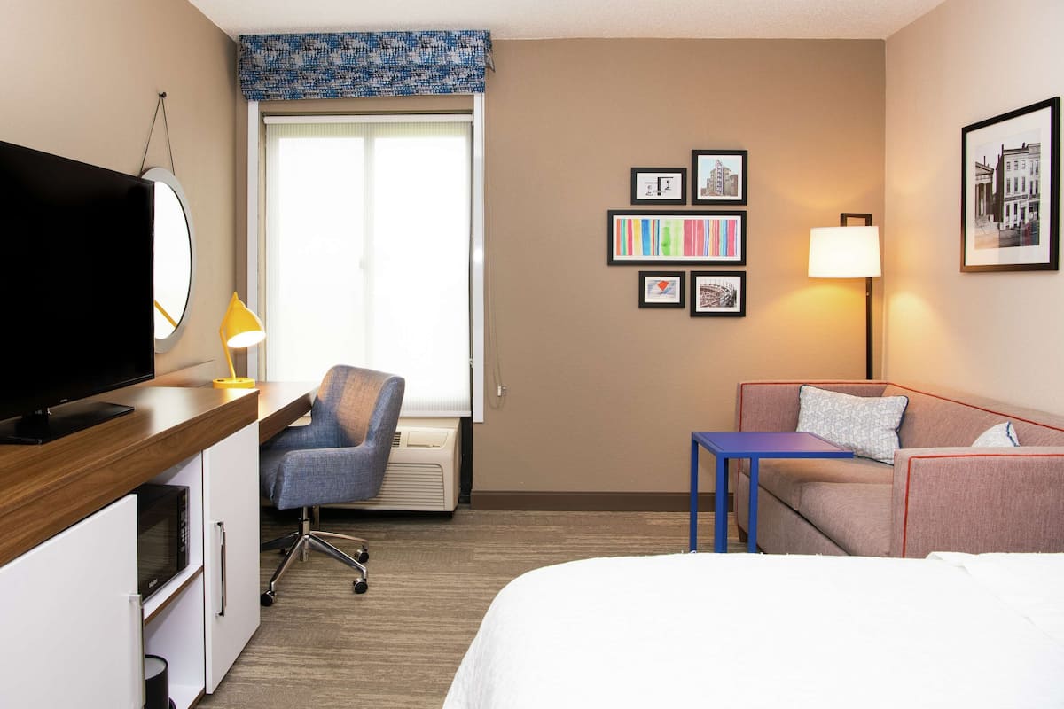A hotel room with a flat-screen TV, a desk with a lamp, a chair, a sofa, a bed, and a window with blinds.