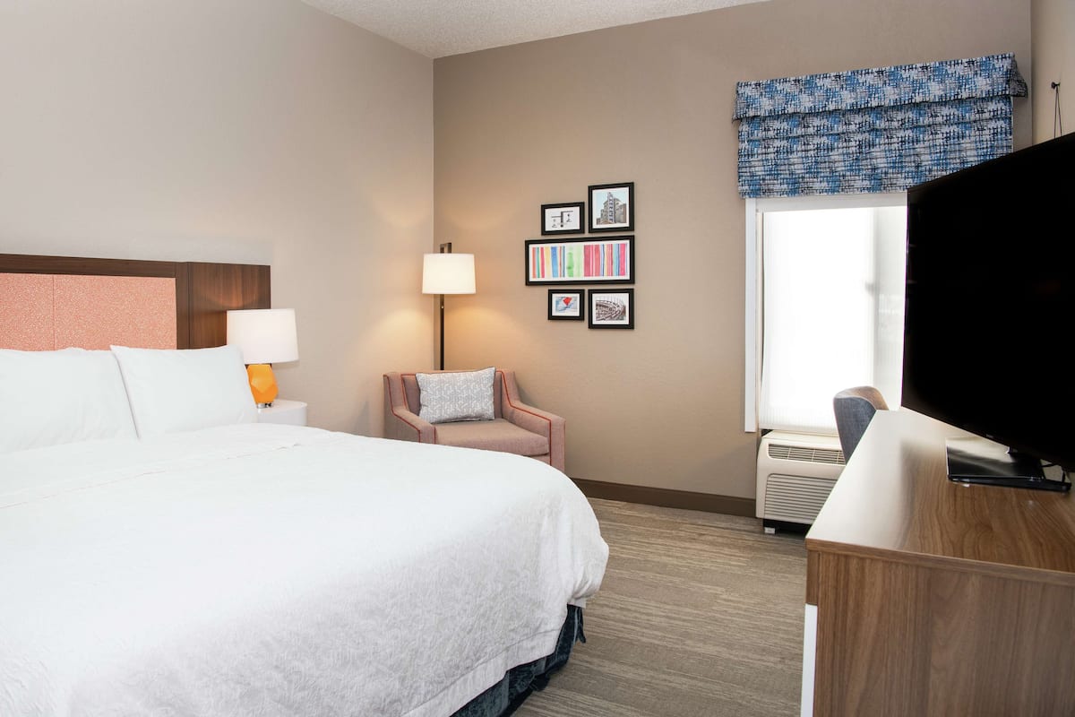 A hotel room with a large bed, a desk, a chair, a lamp, a TV, and framed pictures on the wall.
