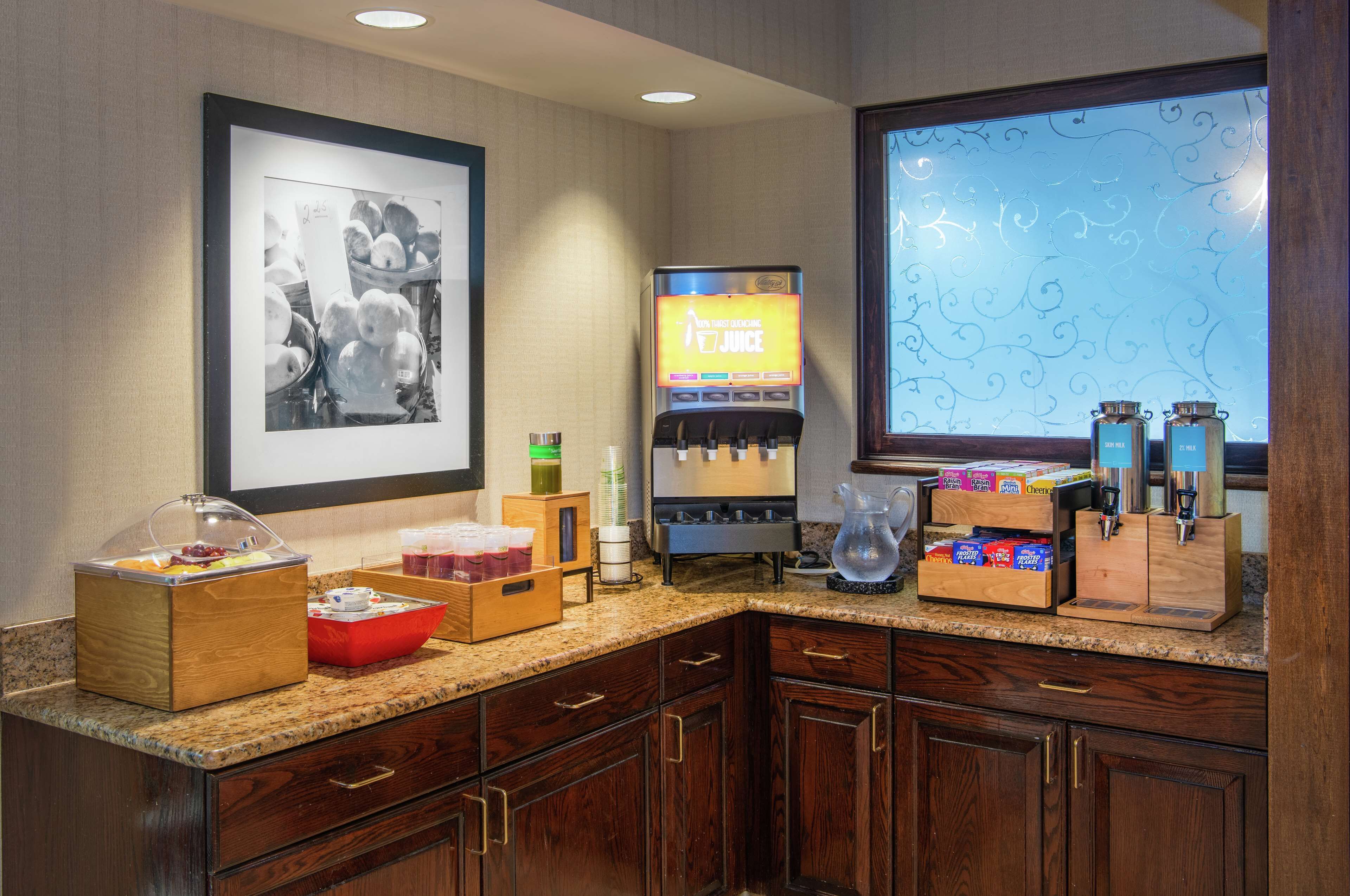 Un espace où est servi le petit-déjeuner à l’hôtel, avec une machine à café, un panier de fruits et divers récipients contenant de la nourriture.