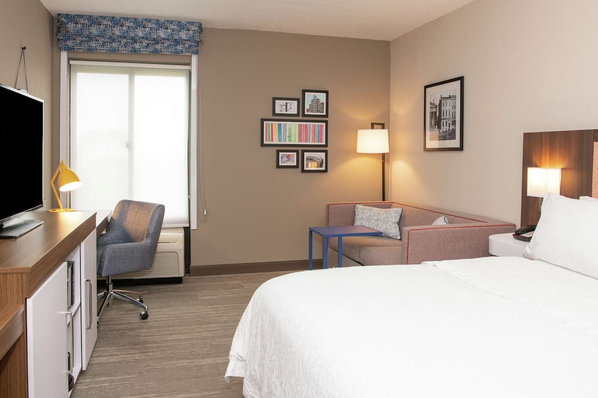 A hotel room with a bed, a desk with a TV, a chair, a sofa, and a window with blinds.