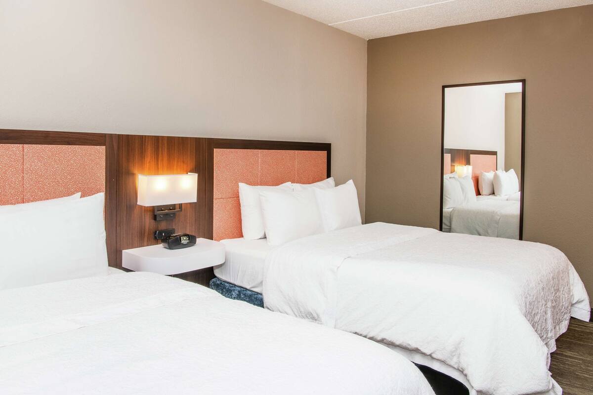 A hotel room with two beds, a nightstand, a lamp, and a mirror.