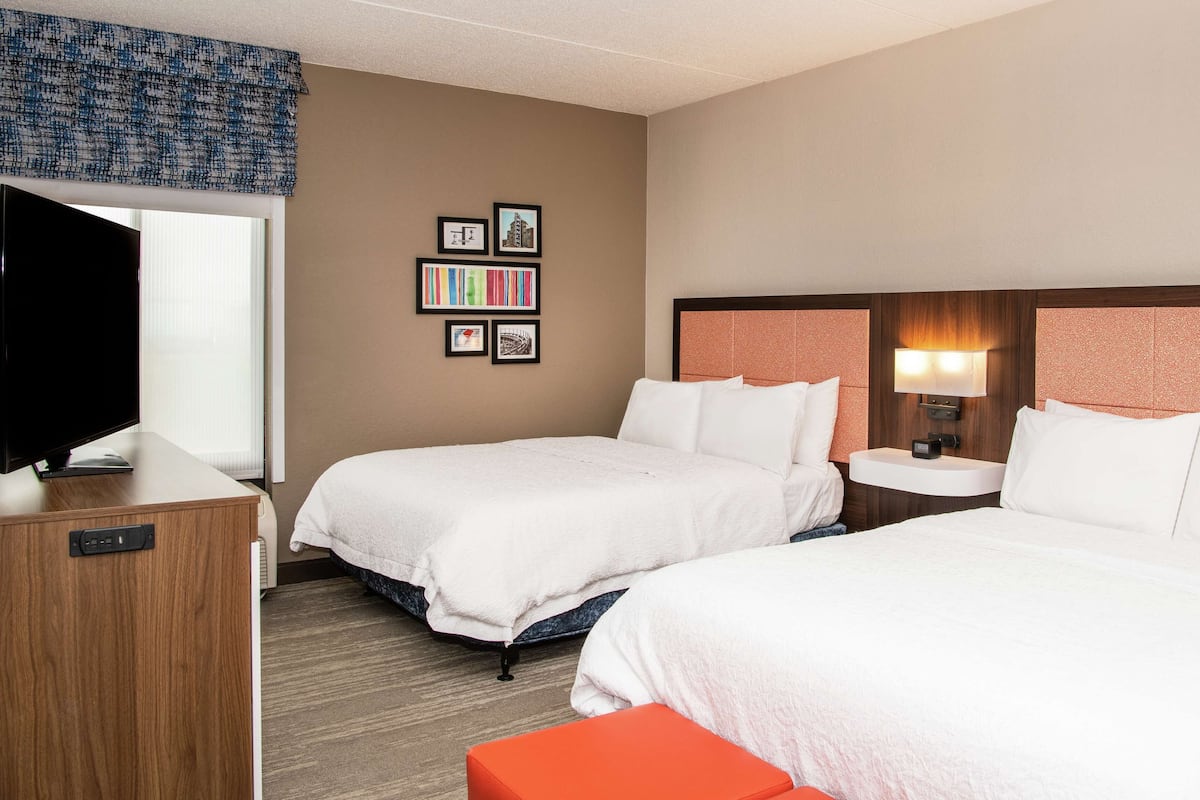 A hotel room with two beds, a TV, a desk, and a wall with framed pictures.
