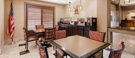 A hotel dining area with a large table, chairs with patterned cushions, a coffee station, and an American flag.
