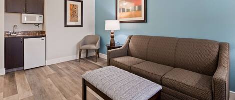 A living room with a brown sofa, a striped ottoman, a small table, a lamp, and a framed artwork on the wall.
