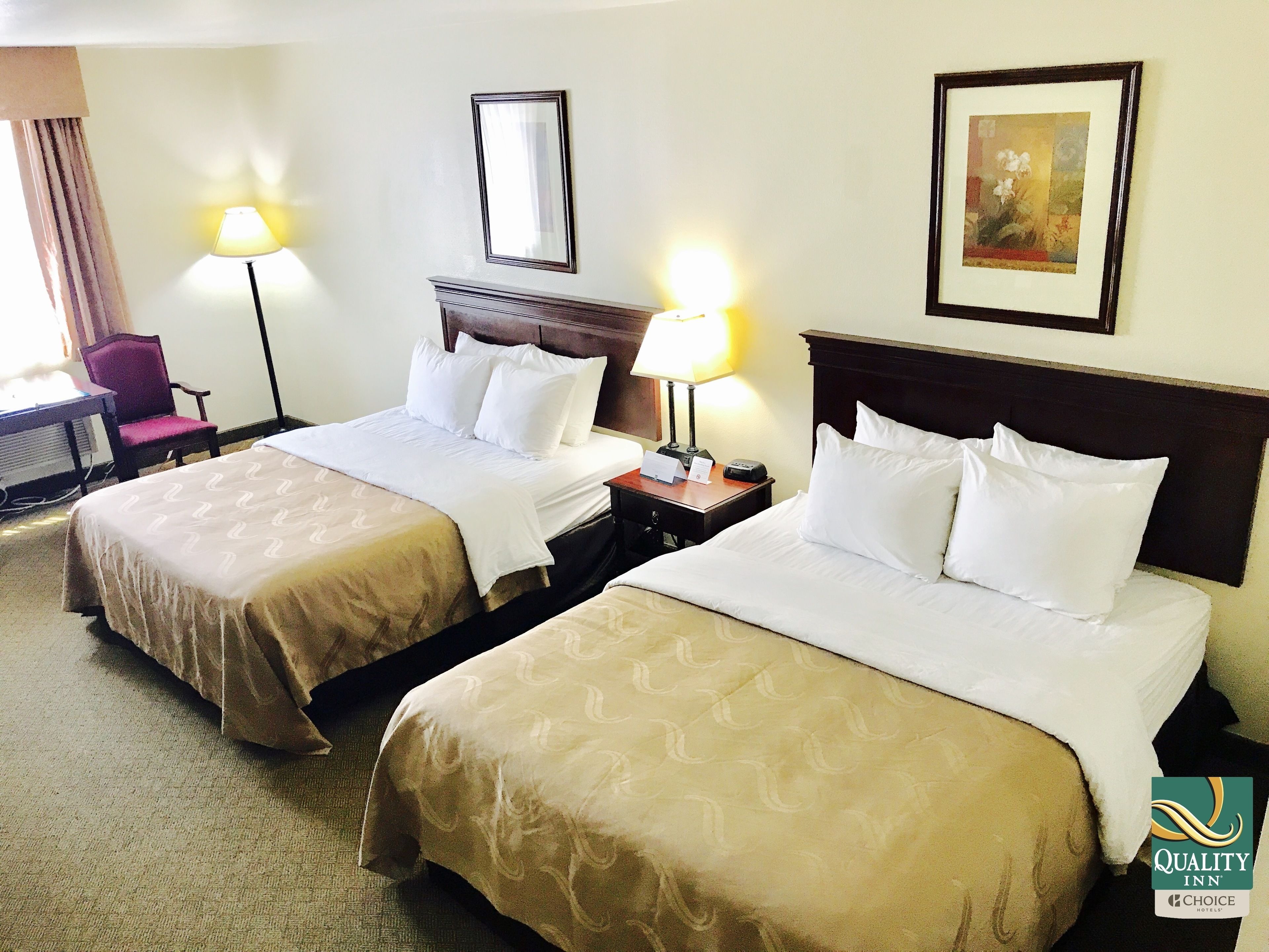A hotel room with two beds, a desk, a chair, and a lamp.
