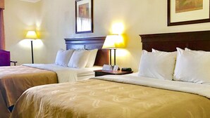 A hotel room with two beds, each with a headboard and a bedside table. There are lamps on both sides of the beds.