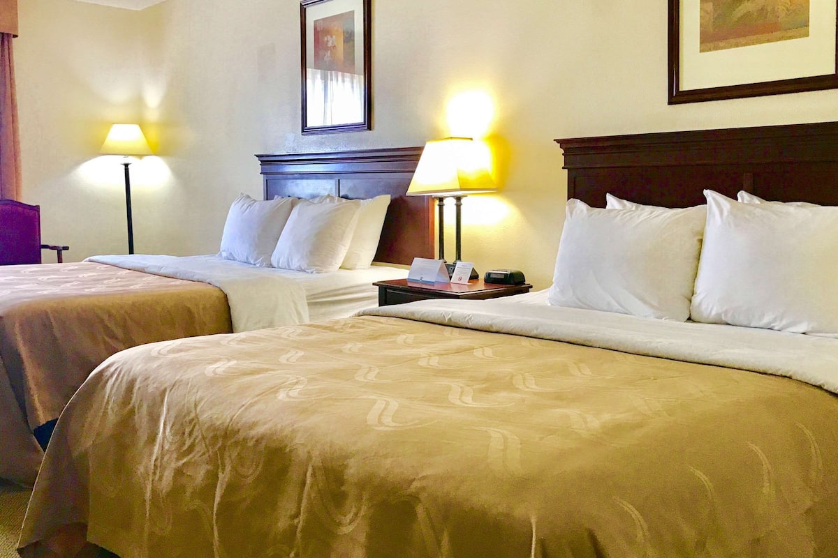 Ein Hotelzimmer mit zwei Betten, jeweils mit Kopfteil und Nachttisch. Auf beiden Seiten der Betten stehen Lampen.