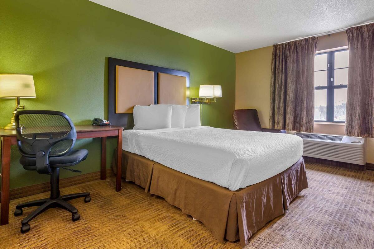 A hotel room with a bed, desk, office chair, lamps, and an air conditioning unit.