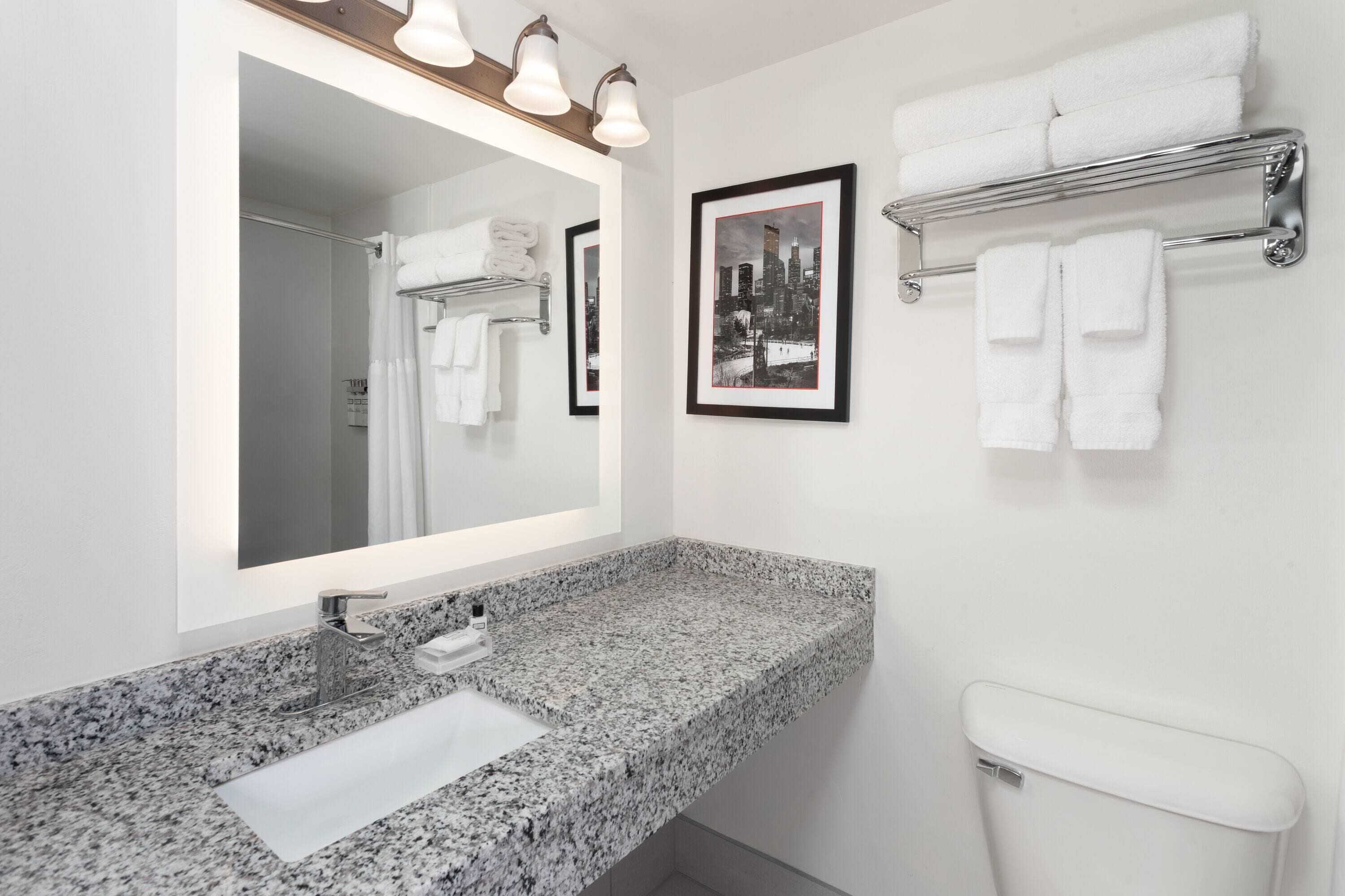 Un baño moderno con lavabo de granito, un espejo grande con iluminación, un cuadro con una vista urbana enmarcada y un perchero con toallas dobladas.