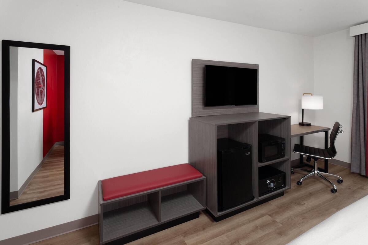 A hotel room with a flat-screen TV mounted on a wooden cabinet, a desk with a lamp, a black office chair, a red bench, and a microwave.