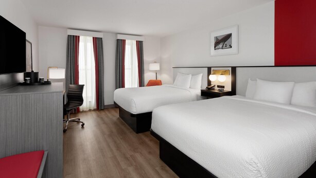 A hotel room with two beds, a desk, a chair, a TV, and a lamp.