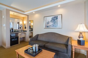 Habitación de hotel con sofá, mesa de centro, lámpara, un cuadro en la pared y baño con lavamanos y espejo.