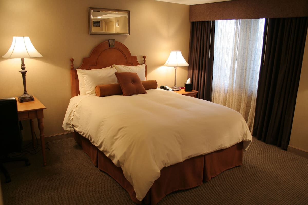 A hotel room with a large bed, two bedside tables with lamps, a desk, and a window with curtains.