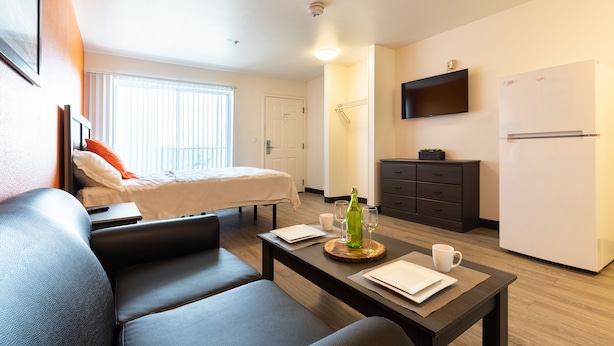 Una habitación de hotel moderna con un sofá, una mesa de comedor con una botella y vasos, una cama, un televisor montado en la pared y un refrigerador.