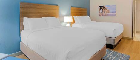 A hotel room with a large bed, a smaller bed, a nightstand with a lamp, a blue wall, and a beach-themed picture.