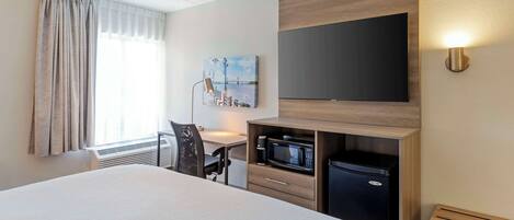 Habitación de hotel con una cama grande, un escritorio con una silla, microondas, refrigerador, televisor de pantalla plana montado en la pared y un cuadro en la pared.