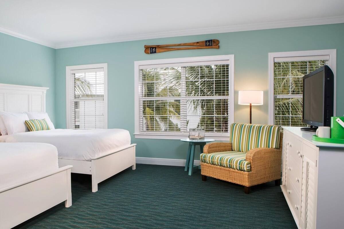 A hotel room with two beds, a small table, a chair, a TV, and a window with a view of palm trees.