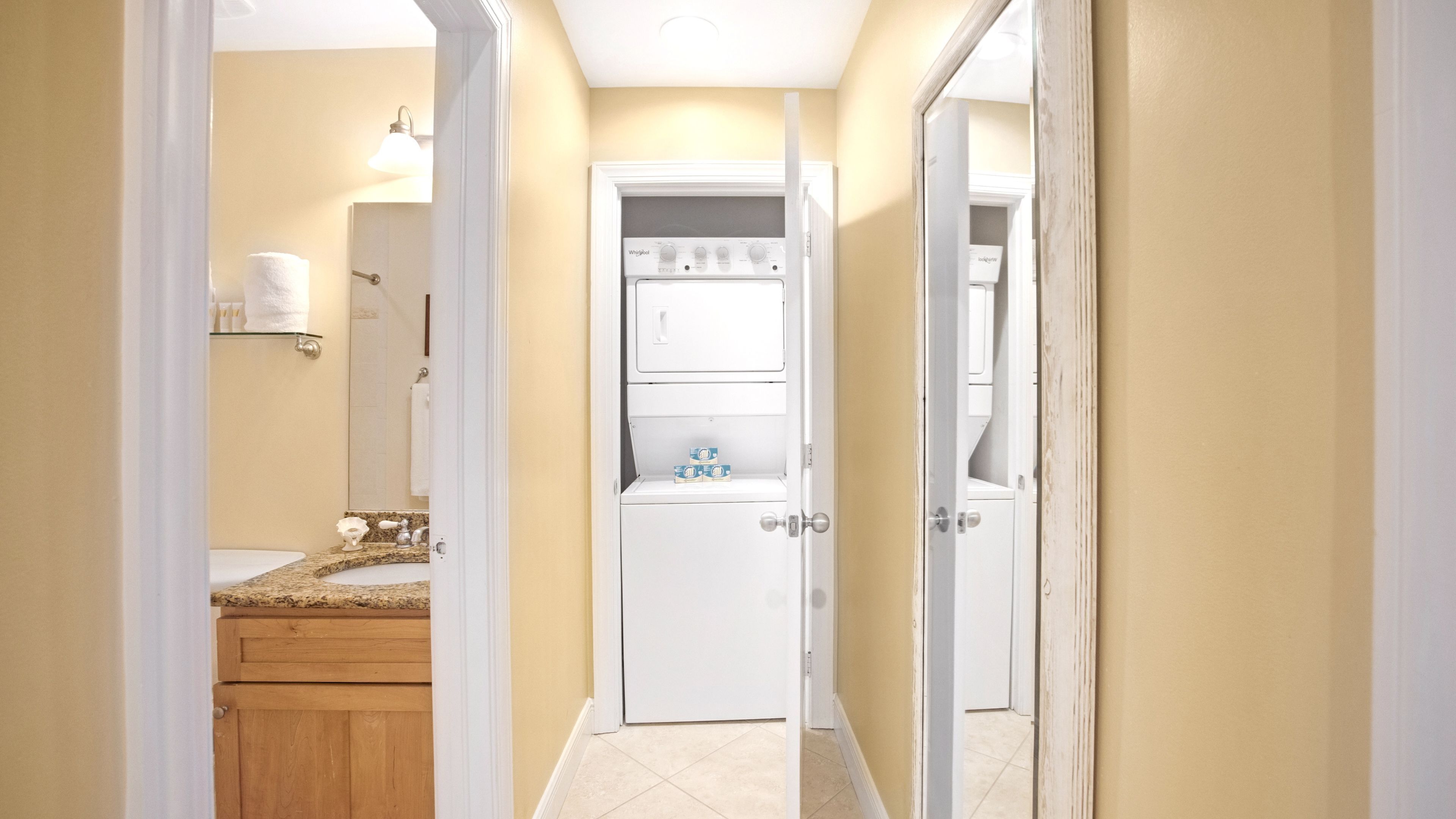 Un couloir étroit, avec une salle de bain sur la gauche, comprenant un lavabo et des toilettes. On peut apercevoir un appareil électroménager blanc dans la salle de bain.