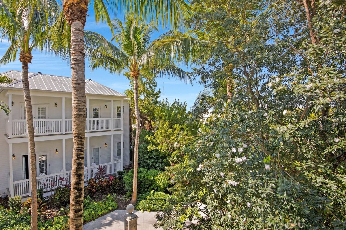 A two-story building with a white exterior, multiple balconies, and surrounded by lush greenery and palm trees.