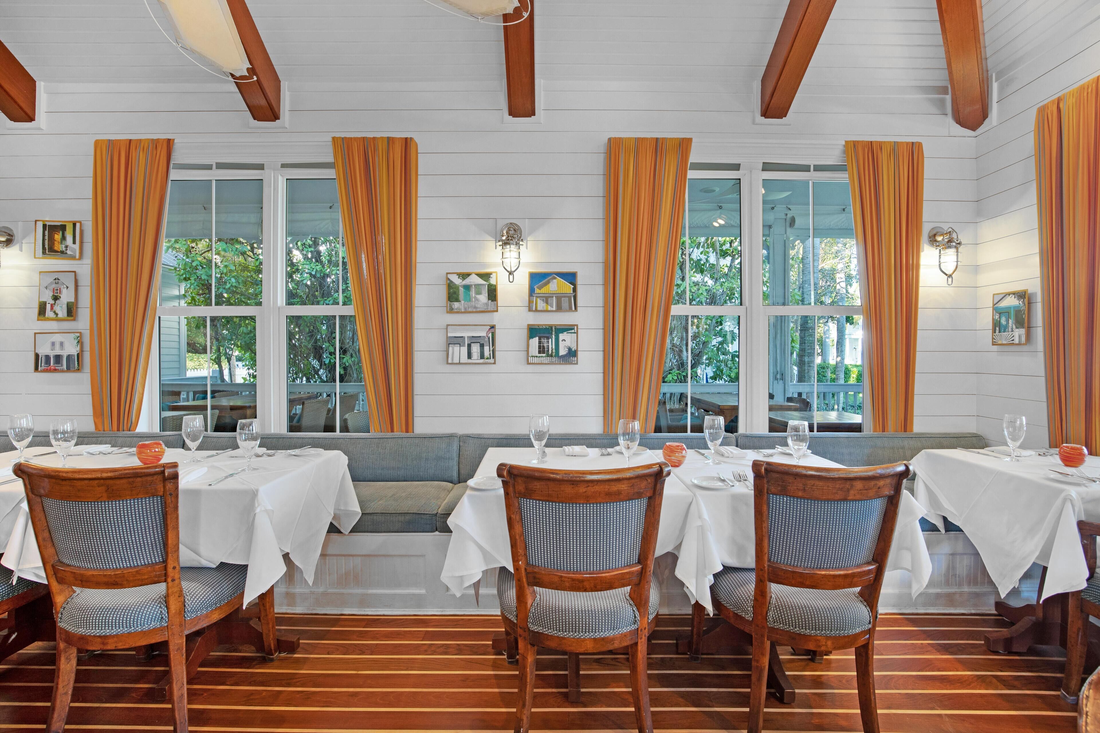 Un comedor con mesas y sillas de madera, manteles blancos y ventanas grandes con cortinas naranjas.