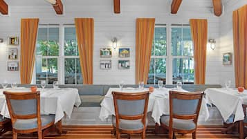 Un comedor con mesas y sillas de madera, manteles blancos y ventanas grandes con cortinas naranjas.