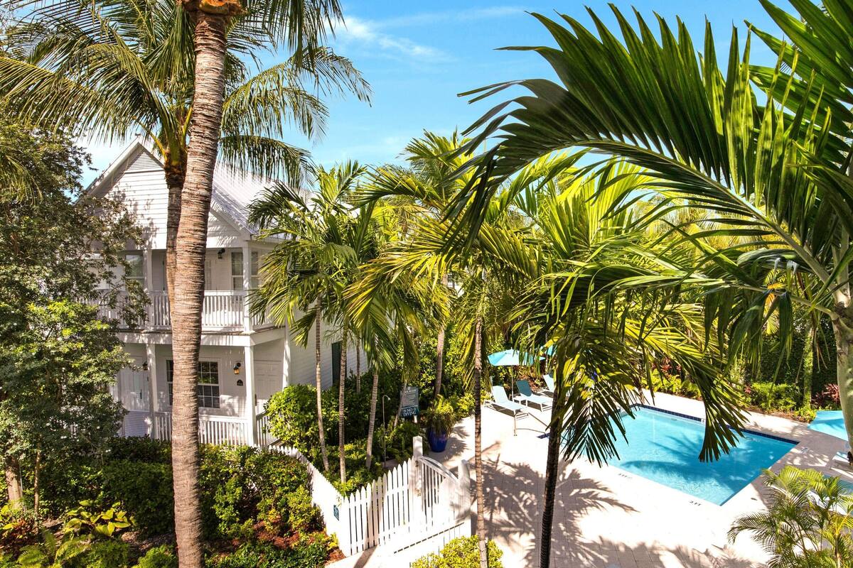 A pool area with palm trees, a white building with balconies, and a clear blue sky.