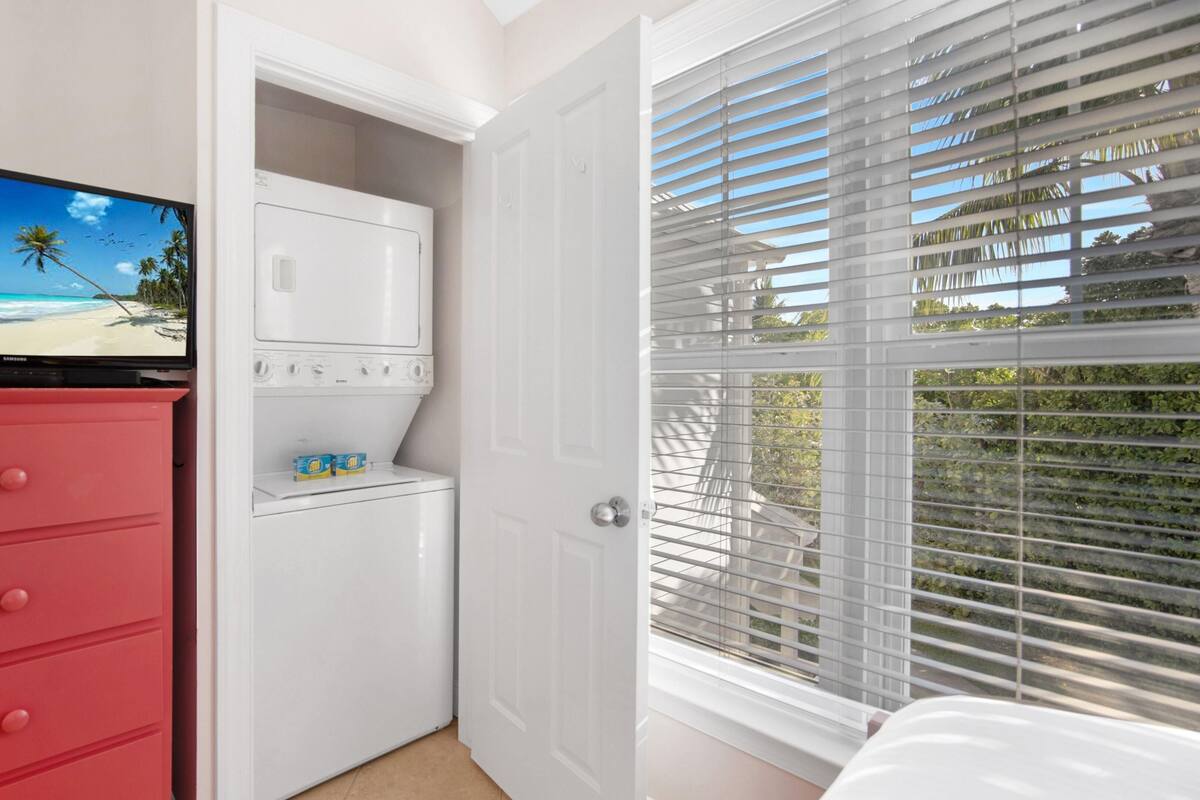 A bedroom with a TV displaying a beach scene, a red dresser, a white wardrobe, and a window with blinds.
