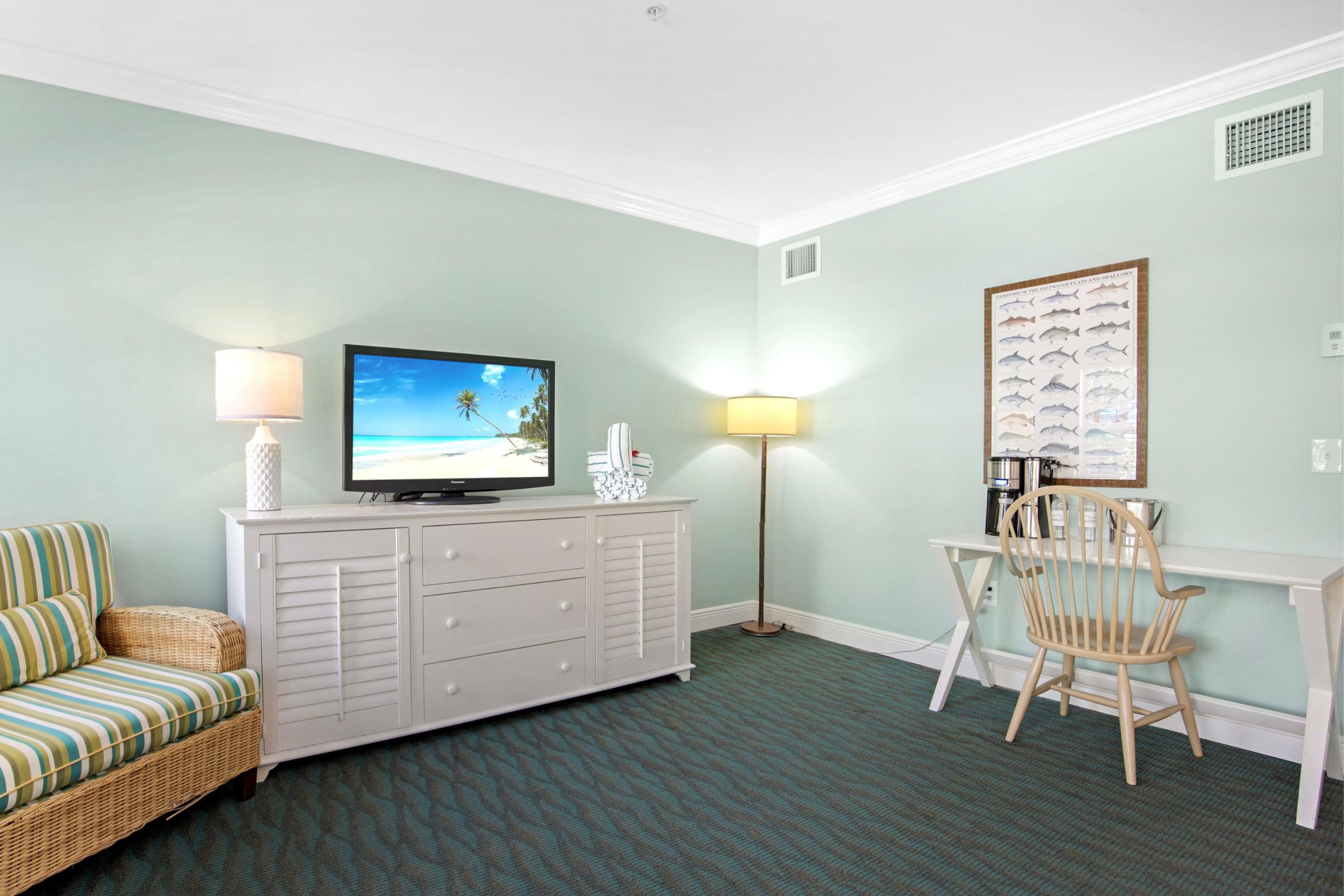 A room with a television on a white dresser, a wicker sofa, a dining table with chairs, and a wall-mounted fish chart.