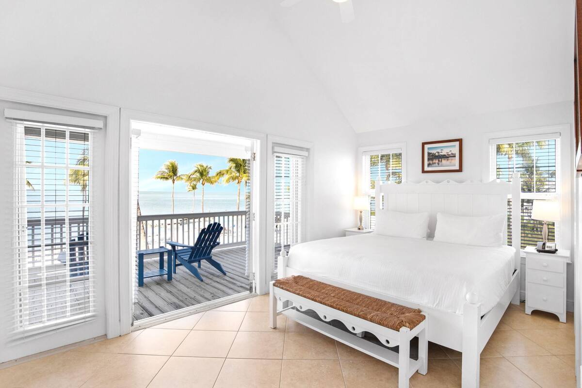 A bedroom with a large bed, a bench, a chair, and a view of the ocean.