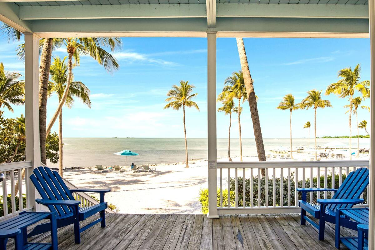 A wooden deck with blue chairs overlooking a beach with palm trees and the ocean.