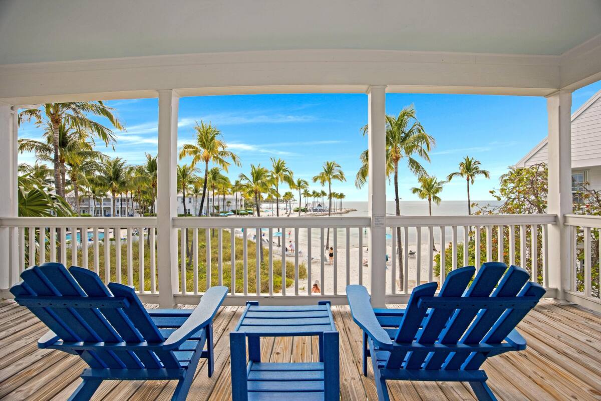A covered outdoor area with blue Adirondack chairs and a wooden table, overlooking a beach with palm trees and a clear blue sky.