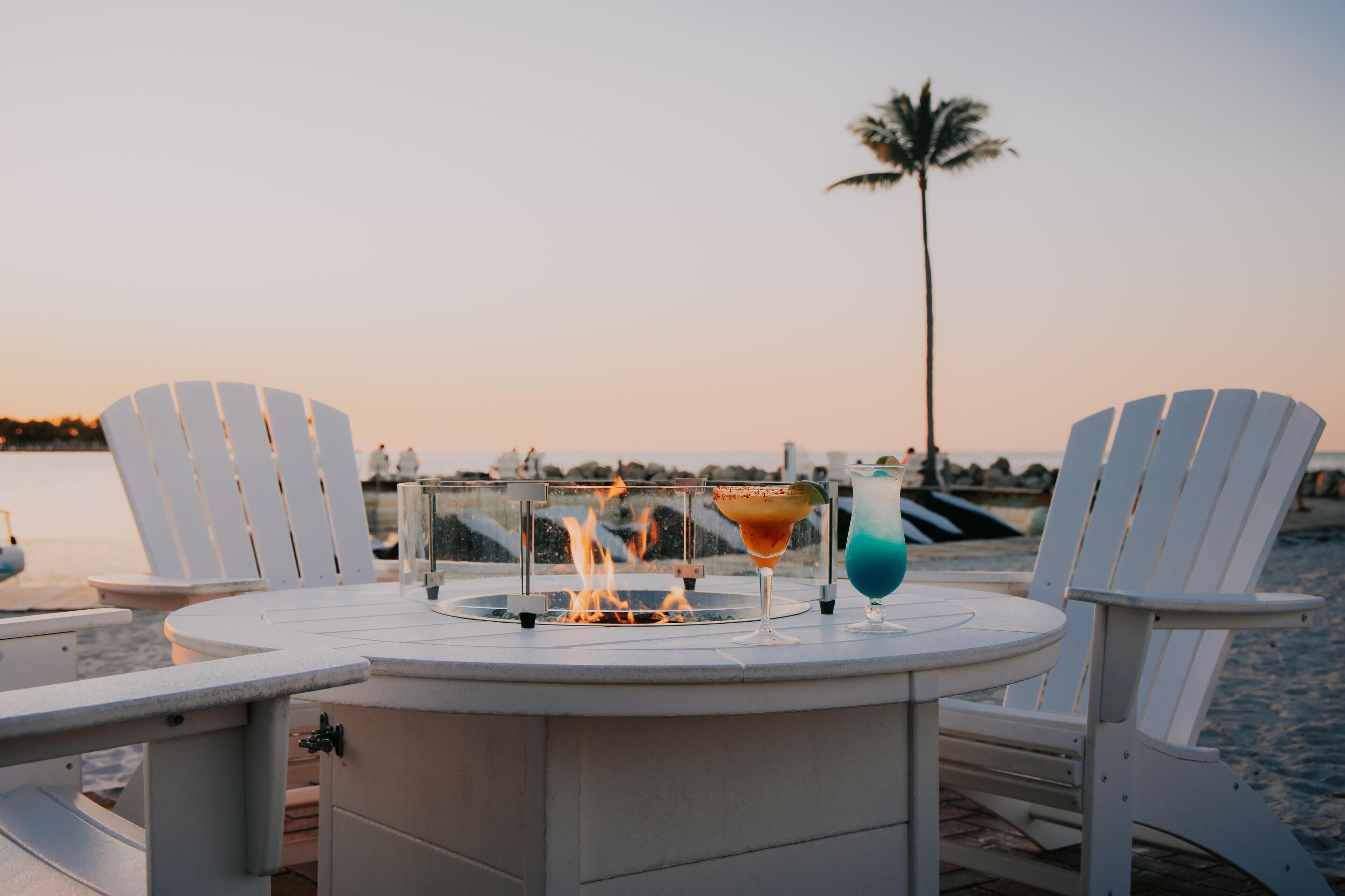 A round outdoor table with a built-in fire feature, two chairs, and two drinks.