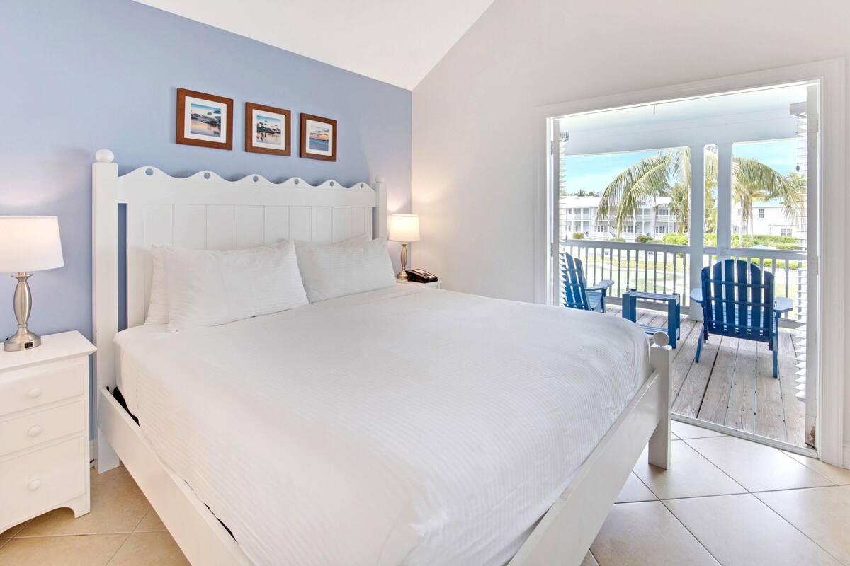 A bedroom with a bed, nightstands, a lamp, and a view of a balcony with chairs.