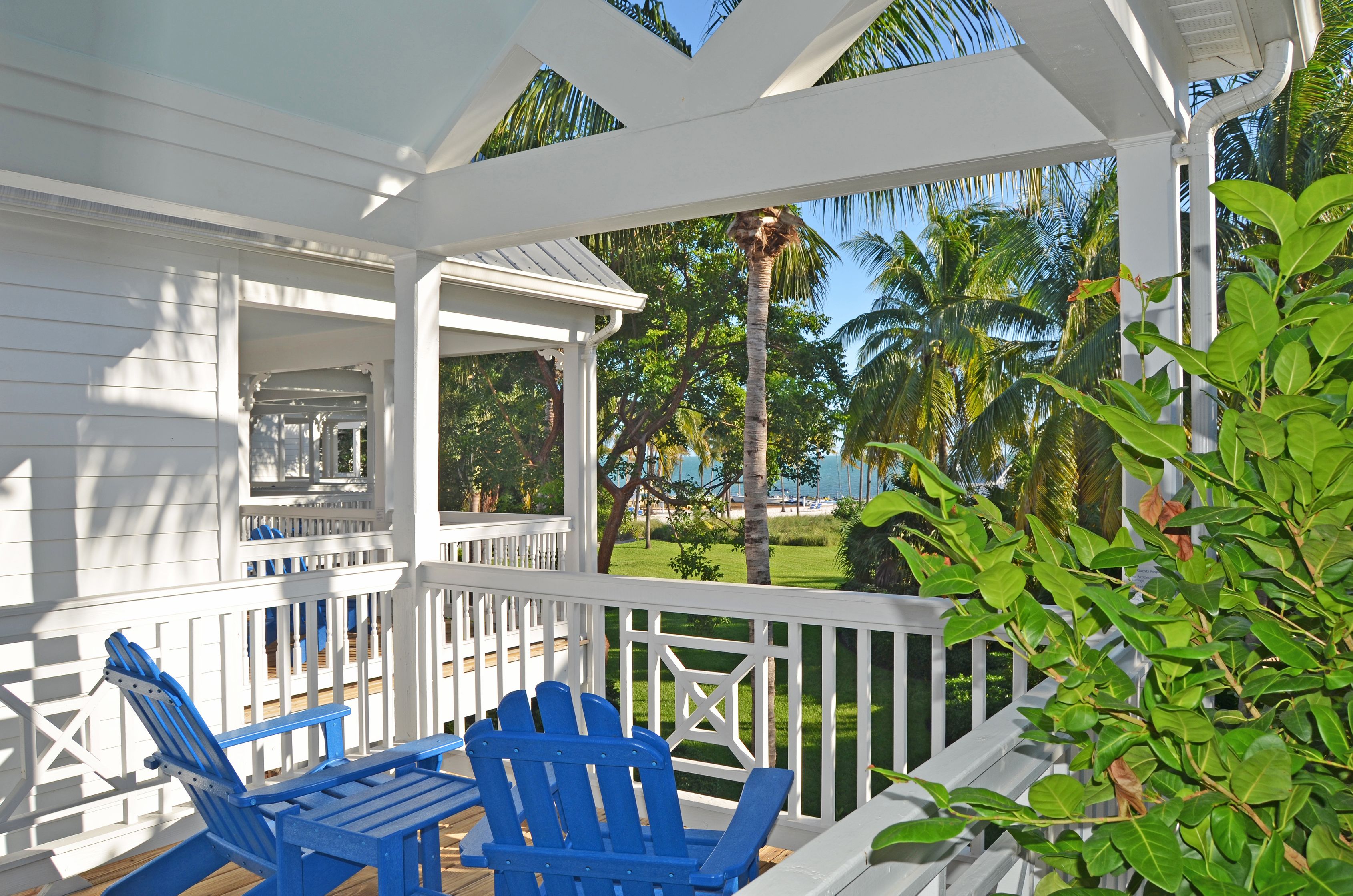 Une terrasse extérieure couverte, dotée de rampes blanches et de chaises Adirondack bleues, qui offre une vue sur une plage et des palmiers.