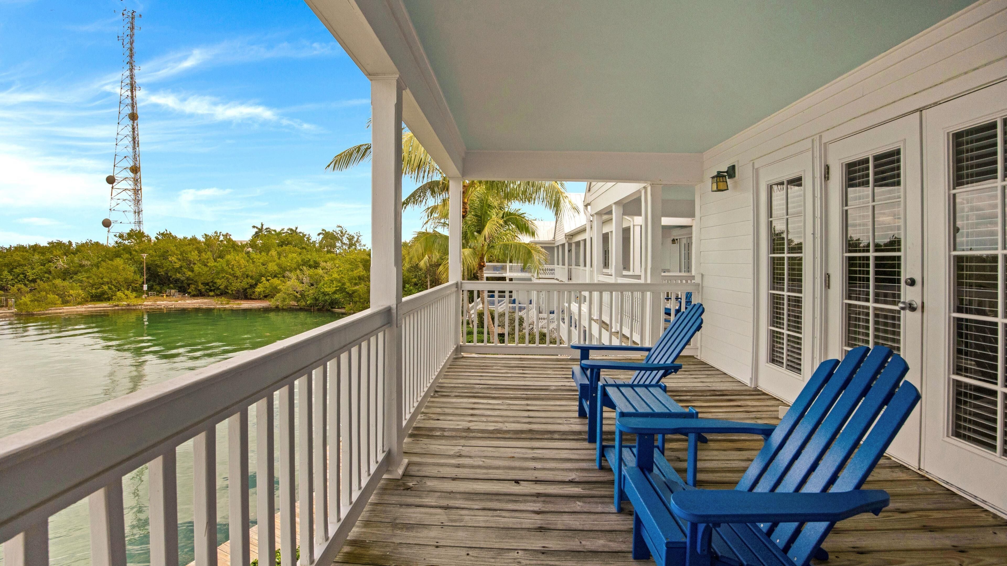 A wooden deck with white railings, blue Adirondack chairs, and a view of the water and trees.